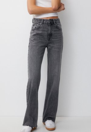 Person, die hoch taillierte, ausgewaschene schwarze Jeans mit geradem Bein, ein weißes kurzes Tanktop und weiße Sneakers vor einem einfarbigen Hintergrund trägt.