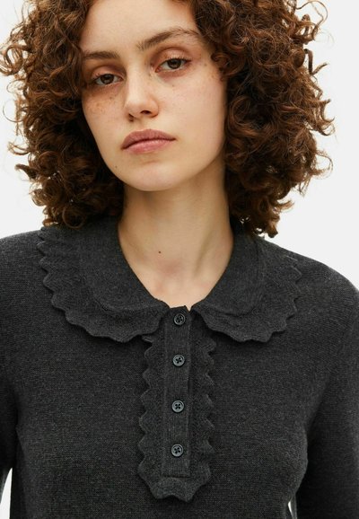 Jonge vrouw met krullend haar, gekleed in een donkergrijze gebreide top met een gegolfde kraag en vier zwarte knopen aan de voorkant.