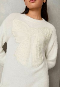 Weißer, flauschiger Pullover mit einem großen, strukturierten Schleifenmotiv auf der Vorderseite, mit feinem Stitching und einer weichen, leicht transparenten Oberfläche.
