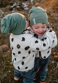 Kaksi lasta, jotka käyttävät vihreitä ja valkoisia raidallisia pipoja ja valkoisia fleecetakkeja, joissa on mustia pilkkuja, halavat toisiaan ulkona.