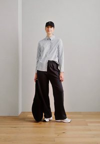 Camicia a righe blu e bianca, pantaloni neri, sneakers bianche e un cappellino nero. In piedi con una borsa texturizzata in mano.