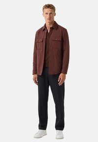 Braune leichte Jacke mit zwei Brusttaschen, schwarzen Knöpfen, über einem braunen Oberteil getragen, kombiniert mit navyblauen Hosen und weißen Sneakers.