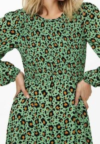 Grünes Kleid mit gerafftem Oberteil, das mit einem schwarz-orangen Leopardenmuster versehen ist. Lange Ärmel mit gepufften Details und einem fließenden Rock.