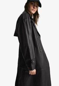 Svart läder trenchcoat med dubbelknäppt knäppning, breda slag och en strukturerad passform. Har detaljerad sömnad och knäppta manschetter.