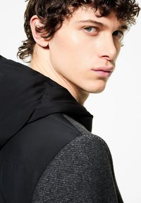 Giacca nera con cappuccio dalla texture liscia, maglione grigio sottostante con un motivo lavorato a maglia. Il modello ha i capelli ricci e occhi blu.