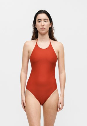 Žena stoji okrenuta prema naprijed, nosi jednodijelni crveni kupaći kostim s visokim ovratnikom na jednostavnoj svijetloj pozadini.