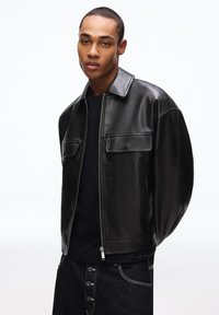 Junger Mann trägt eine schwarze Lederjacke über einem schwarzen Hemd und dunkle Jeans mit Knopfleiste vor weißem Hintergrund.