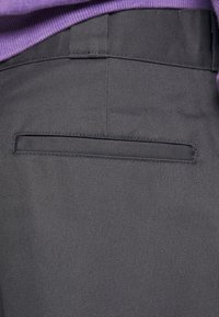 Gros plan sur un pantalon en tissu noir avec une poche passepoilée à l'arrière et une ceinture côtelée violette visible en haut.
