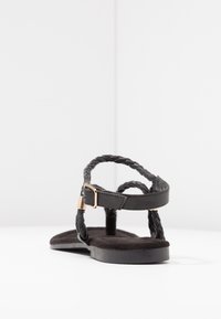 Black sandal with braided leather straps, flat suede footbed, and gold-tone hardware buckle. Soft texture and minimalistic design.