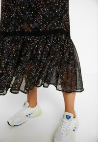 Robe noire à fleurs avec un ourlet froncé, présentant des motifs de fleurs multicolores et des détails en dentelle. Associée à des baskets blanches avec des accents bleus.