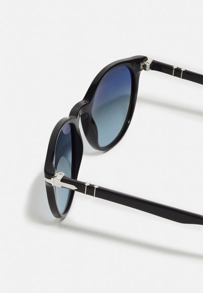 Lunettes noires avec monture ronde, verres dégradés bleus, accents métalliques sur les branches et texture plastique lisse.