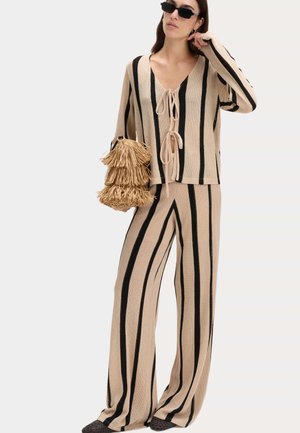 Femme portant un haut en maille rayé vertical beige et noir et un pantalon à jambes larges, tenant un sac beige à franges et portant des lunettes de soleil noires.