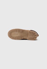 Gros plan de la semelle en caoutchouc marron d'une chaussure décontractée avec des crampons profonds et le logo "Wrangler" en relief, sur un fond clair.