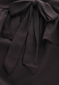 Sötét szürke blúz texturált anyagból, fodros kialakítással és nagy masnival a deréknál, hosszú, lelógó szalagokkal.