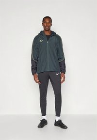 Donkergroene jas met rits, zwarte mouwen en gele accenten, gecombineerd met zwart aansluitende broeken en zwarte sportschoenen, staand op een effen achtergrond.