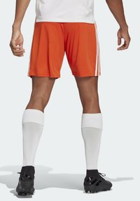 Shorts de sport orange avec des bandes blanches sur les côtés, assortis à des chaussettes blanches montant jusqu'aux genoux et des chaussures de football noires. Design simple, matériau léger.