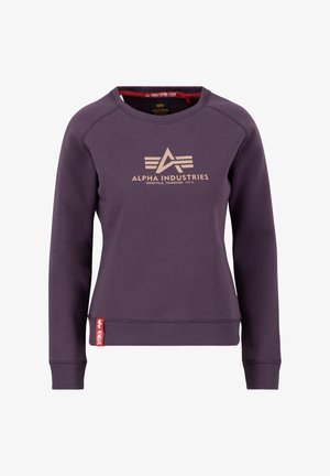Purpurna majica s crewneck iz mehke tkanine, ki na sprednji strani vsebuje zlati logotip in rdečo oznako na levem robu. Srednje prileganje z rebrastimi manšetami.