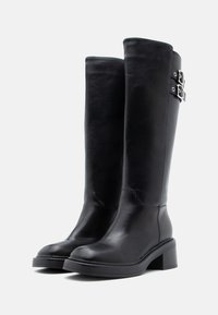 Μαύρες δερμάτινες μπότες μέχρι το γόνατο με λείο φινίρισμα, που διαθέτουν δύο ασημί αγκράφες στις πλευρές και χοντρό τετράγωνο τακούνι.