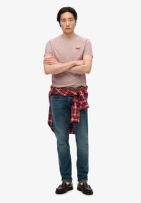 Camiseta a rayas rojas y blancas, jeans azules, camisa de cuadros atada a la cintura, cinturón marrón y zapatos oscuros, de pie con los brazos cruzados.