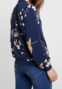 Námořnická bomberová bunda s květinovým potiskem v krémové a béžové barvě, žebrované manžety a pás. Měkká látka a volný střih, vhodná pro každodenní nošení.