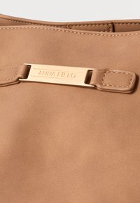 Syntetická kabelka v hnedej farbe s hladkou textúrou, zlatým kovaním a značkou "ANNA FIELD" na obdĺžnikovej platničke.