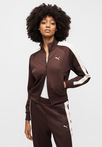 Καφέ ζακέτα φόρμας με ψηλό γιακά, φερμουάρ μπροστά και κρεμ ρίγες στα πλαϊνά. Διαθέτει λογότυπο Puma στο στήθος και τσέπες στα πλευρά.