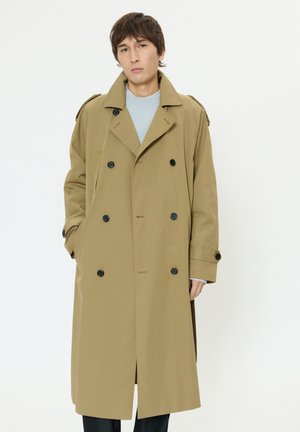 Beiger, doppelseitiger Trenchcoat mit schwarzen Knöpfen, Schulterklappen und einer knielangen Länge. Getragen über einem hellblauen Pullover.