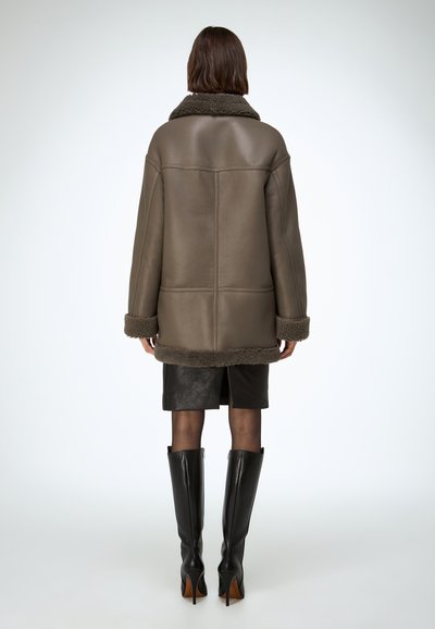 Abrigo marrón de cuero con cuello de piel, hombreras caídas y corte recto, combinado con una minifalda negra de cuero y botas altas negras.