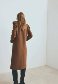 Bruine wollen jas met een gestructureerd silhouet, lange mouwen en een rechte snit, gecombineerd met zwarte laarzen tot aan de knie, tegen een lichte achtergrond.