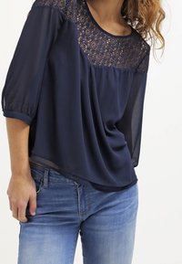 Blusa blu navy con pannello in pizzo nella parte superiore, materiale semi-trasparente e maniche tre quarti, abbinata a jeans azzurri.