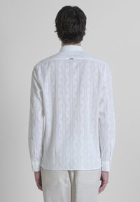 Camisa blanca de mangas largas con un patrón geométrico texturizado. Cuello rígido y puños con botón, con un sutil brillo en la tela.