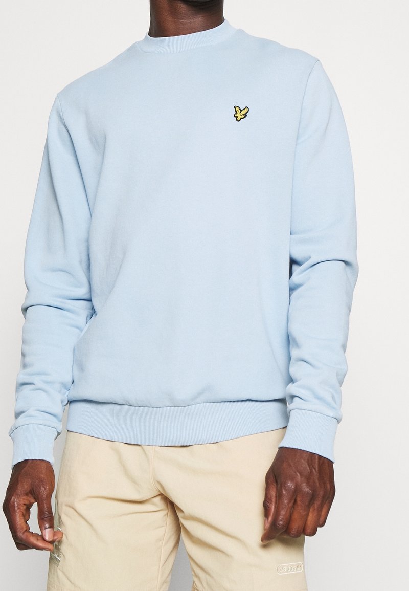 Homme portant un sweat-shirt bleu clair à encolure ronde avec petit logo d'oiseau jaune sur la poitrine et pantalon beige, sur fond uni.