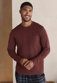 Braunes Langarmhemd mit einer Brusttasche, aus weichem Stoff. Das Hemd wird mit schwarz-grau karierten Hosen kombiniert, die einen lockeren Schnitt haben.