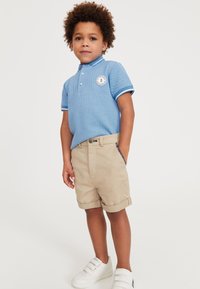Blauwe poloshirt met een textuurpatroon en witte accenten, gecombineerd met kaki shorts. Accessoires omvatten witte instappers.