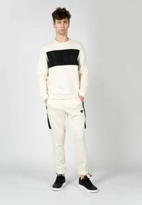 Sudadera crema con un panel horizontal negro, combinada con joggers crema que tienen bolsillos laterales negros y zapatillas negras.