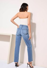 Jeans alta de cintura en azul claro con pernera recta, bolsillos traseros y un acabado desgastado, combinados con una blusa blanca sin tirantes y tacones negros.