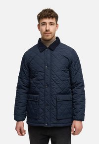 Chaqueta acolchada azul marino con cuello de pana, dos bolsillos frontales y un acabado texturizado. Diseño de cierre con botones; forma rectangular en general.