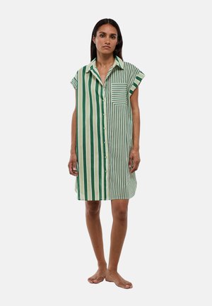 Femme debout pieds nus portant une robe chemise boutonnée à manches courtes avec des rayures verticales vertes et blanches et une poche poitrine rayée.