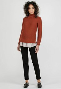 Rust-colored ribbed turtleneck sweater with side slits, layered over a white striped shirt, paired with black slim-fit pants and black shoes.