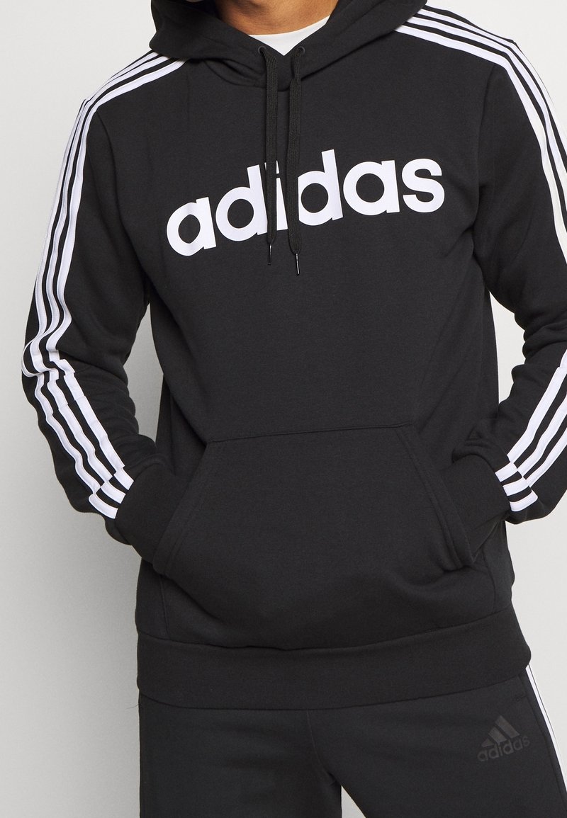 Personne portant un sweat à capuche noir Adidas avec des bandes blanches sur les manches et le logo Adidas à l'avant, les mains dans la poche avant.
