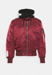 Giacca bomber in satin bordeaux con cappuccio nero, chiusura con zip, polsini a costine e tasca sulla manica sinistra. Presenta dettagli a strisce e un patch con logo.