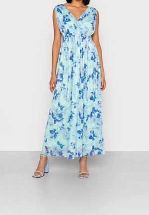Kobieta ubrana w niebiesko-białą kwiecistą sukienkę bez rękawów, z dekoltem w serek, sięgającą do ziemi, w połączeniu z jasnoniebieskimi sandałami na słupku z odkrytymi palcami.