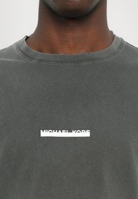 Pilkas medvilnės marškinėliai, su apvaliu kaklu ir trumpomis rankovėmis, su horizontaliai spausdintu baltu "MICHAEL KORS" logotipu ant krūtinės.