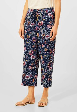 Frau trägt marineblaue, gekürzte Hosen mit einem bunten Blumenmuster, kombiniert mit beigen Riemchensandalen flach.