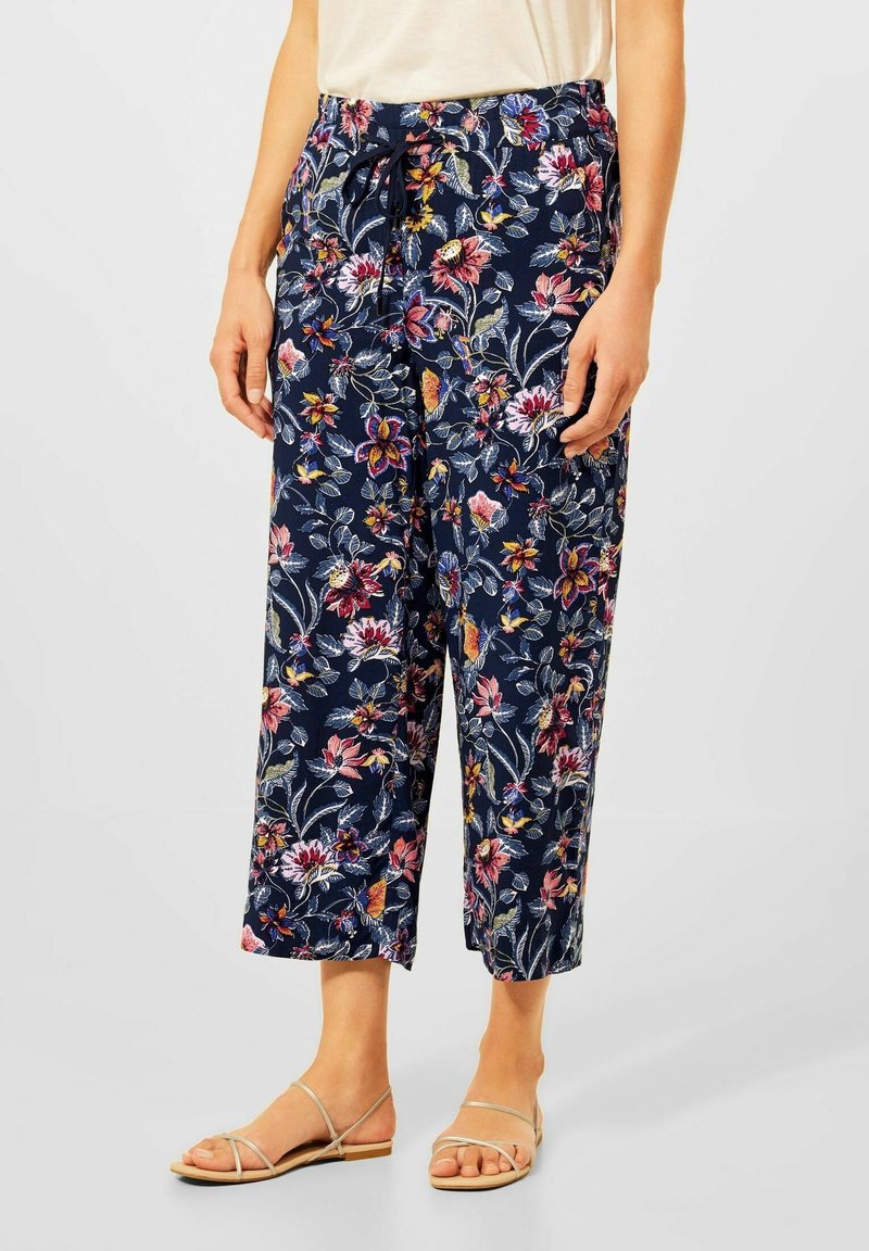 Femme portant un pantalon court bleu marine avec un motif floral coloré, associé à des sandales plates beiges à lanières.