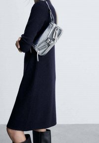 Sac à main argent métallisé avec des fermetures éclair et des accents en cuir, porté avec une robe bleu marine, assorti à des bottes noires montantes jusqu'aux genoux.