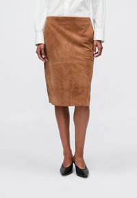 Bruine suède kokerrok tot de knie, gedragen met zwarte puntige schoenen en een witte blouse met lange mouwen, staande tegen een effen achtergrond.
