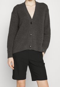Grå stickad cardigan med djup V-hals, fem knappar framtill och ribbat mönster. Kombinerad med svarta shorts.