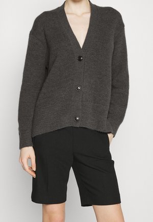 Cárdigan gris de punto con un escote en V profundo, cinco botones en el frente y textura acanalada. Combinado con pantalones cortos negros.