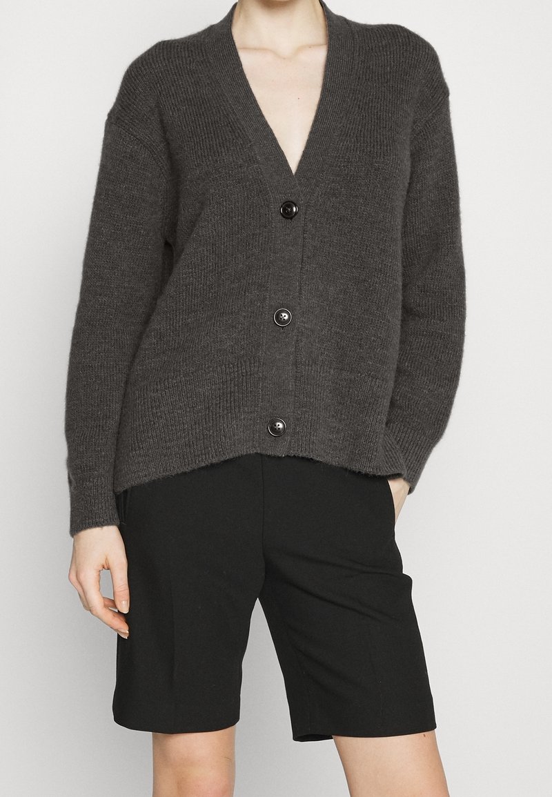 Grå stickad cardigan med djup V-hals, fem knappar framtill och ribbat mönster. Kombinerad med svarta shorts.